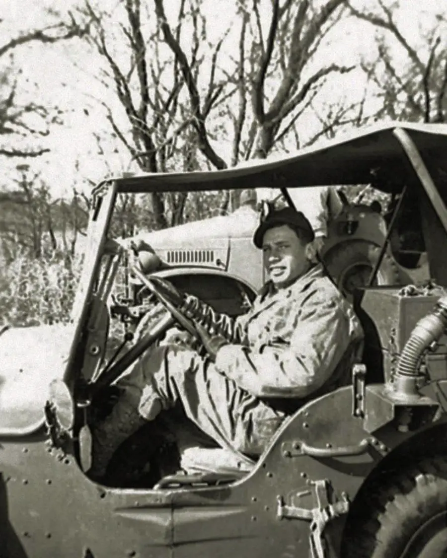 A smiling man in military attire sits in the driver’s seat of a vintage jeep, parked outdoors with leafless trees in the background.