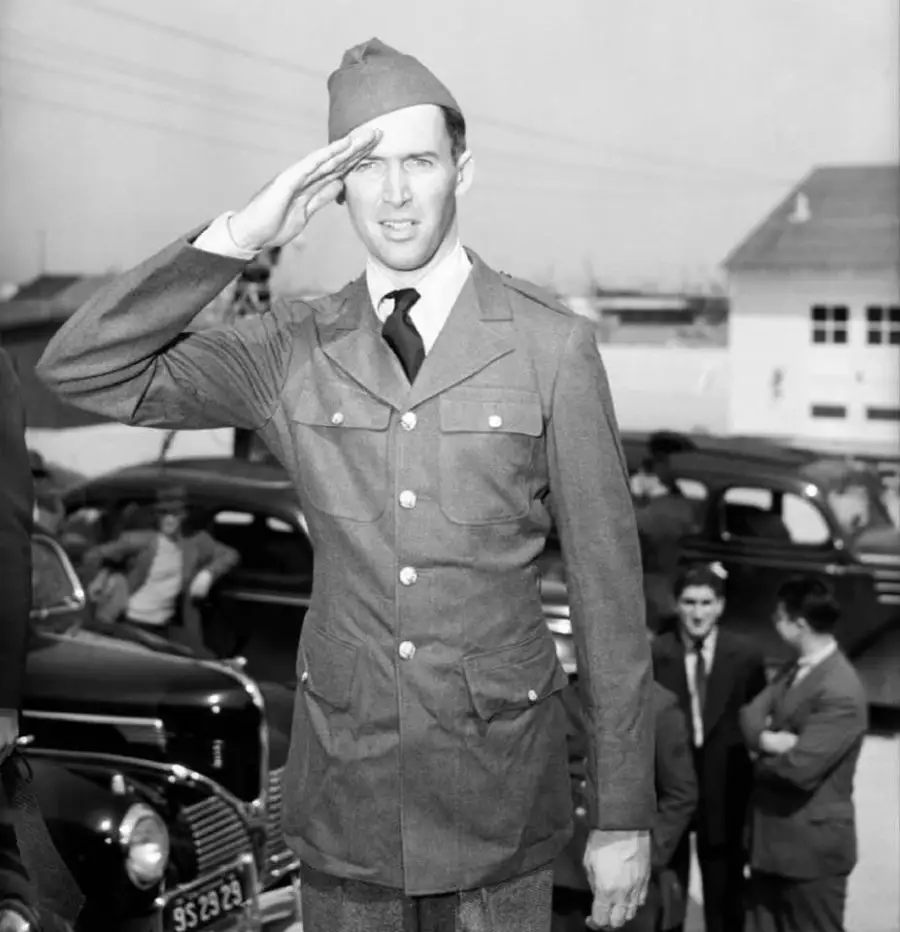 A man in a military uniform and cap salutes outdoors, with classic cars and people in suits visible in the background.