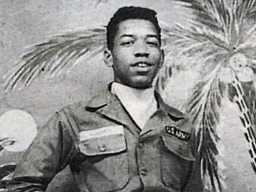 A young Black man in a U.S. Army uniform poses in front of a painted backdrop featuring palm trees. The image is black and white.
