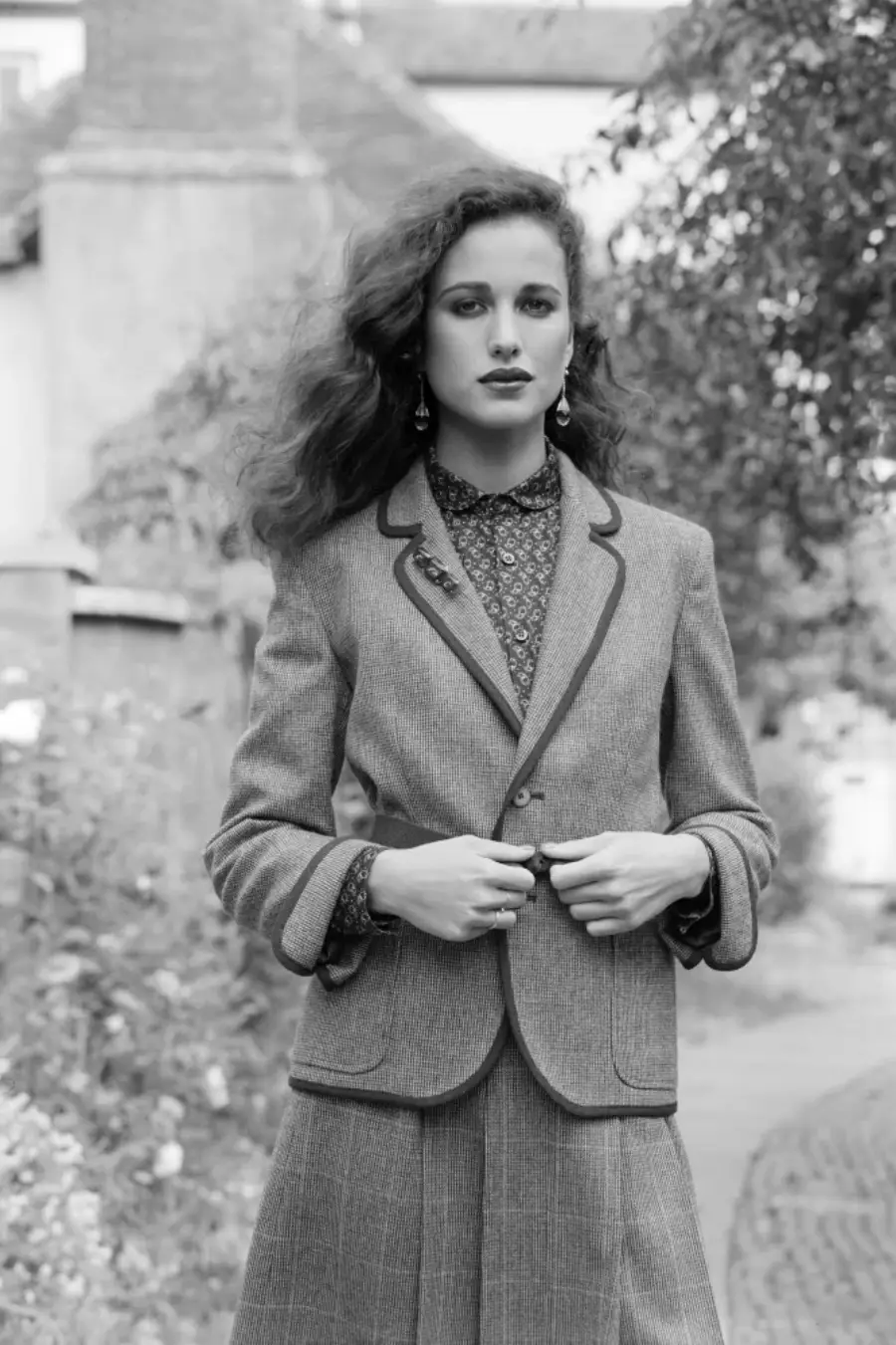 A woman with wavy hair stands outdoors, wearing a tailored blazer over a patterned shirt and a matching skirt. She looks directly at the camera with a confident expression. The image is in black and white.