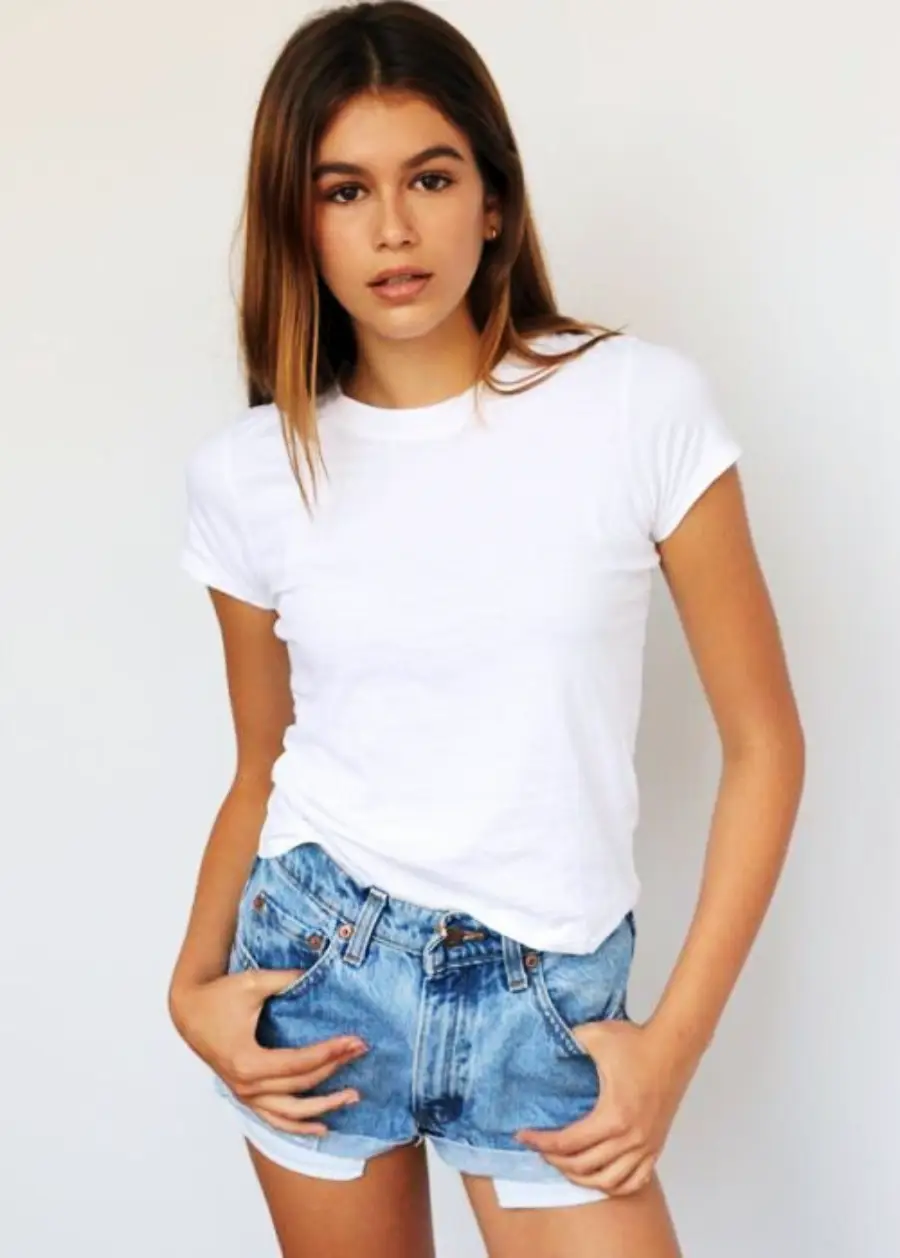 A young woman with long brown hair stands against a plain white background, wearing a white t-shirt and denim shorts. She looks at the camera with a neutral expression and has her hands in her pockets.