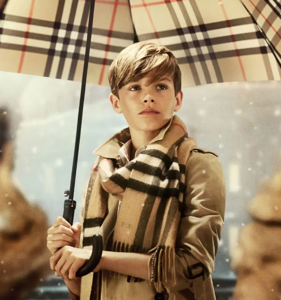 A young boy in a beige trench coat and plaid scarf stands under a matching plaid umbrella, looking to the side. Snow falls around him, and the background is softly blurred.