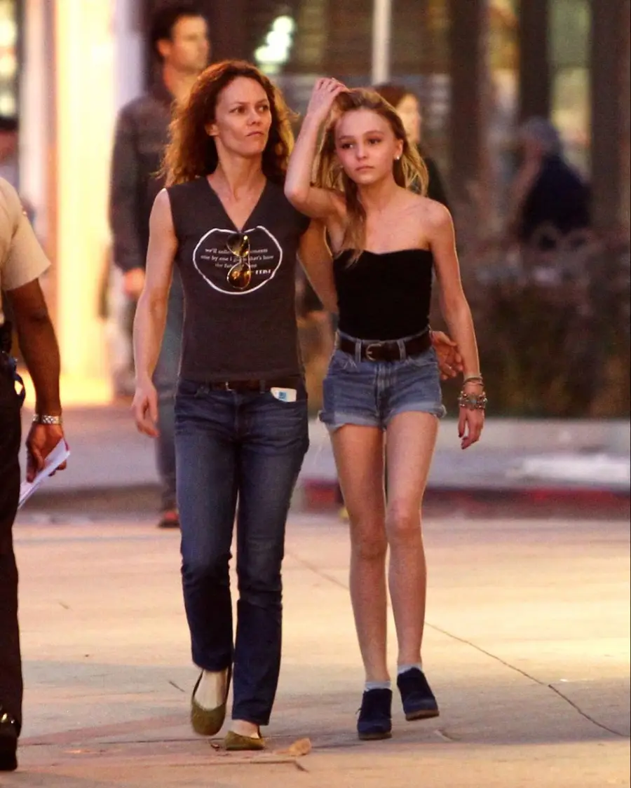 A woman and a teenage girl walk side by side on a city sidewalk in the evening; the woman has her arm around the girl's shoulders as they both look ahead.