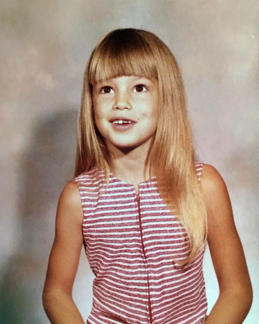 A young girl with long blonde hair and bangs smiles while looking slightly upward. She wears a sleeveless red and white striped top and poses against a neutral, softly textured background.
