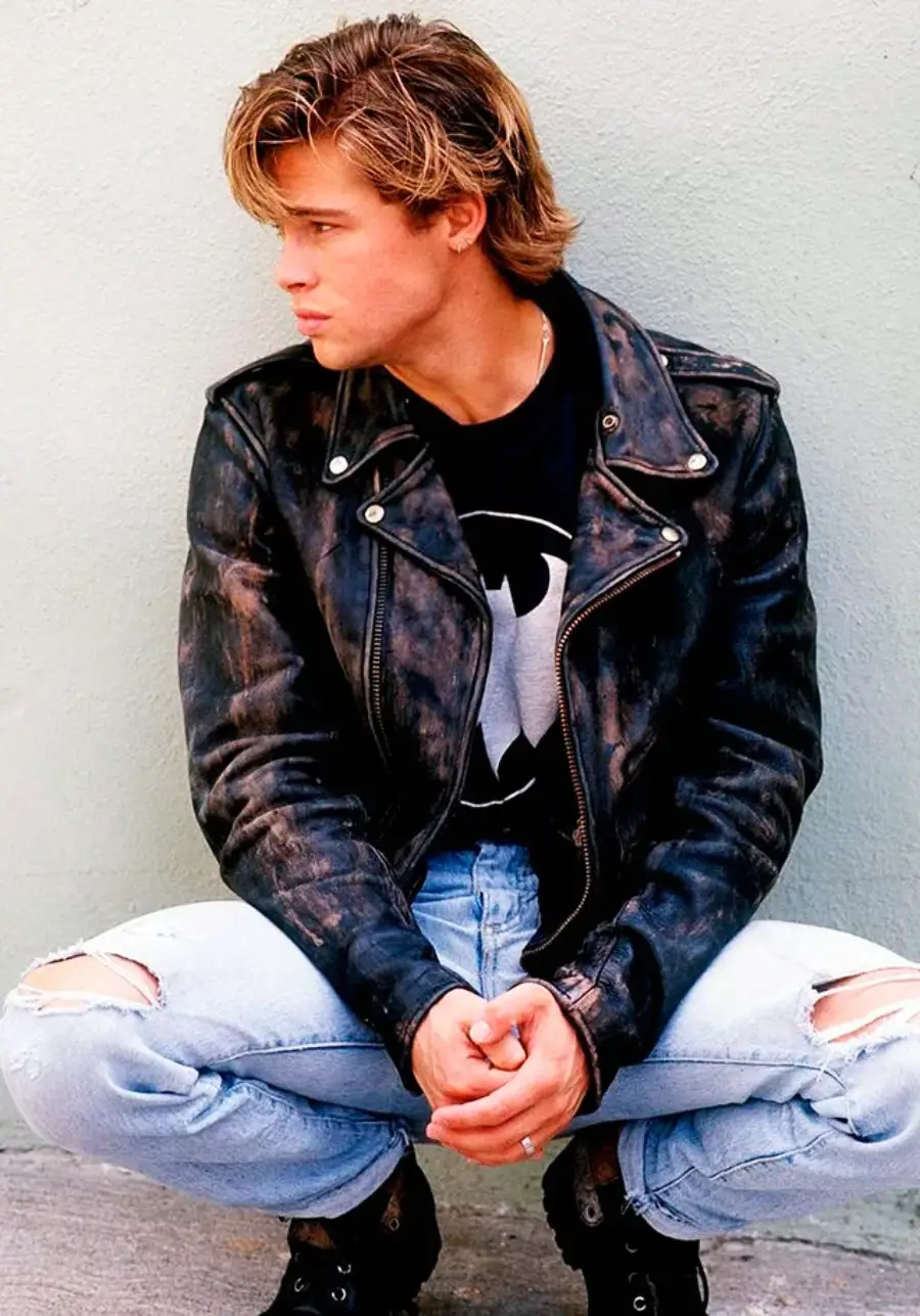 Young man with blonde hair sits on his heels, wearing a black leather jacket, ripped light blue jeans, and a black T-shirt with a Batman logo, looking to his left in front of a plain light-colored wall.