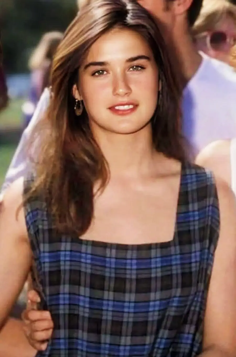 A young woman with long brown hair wears a blue and gray plaid sleeveless dress, smiling outdoors with people blurred in the background.