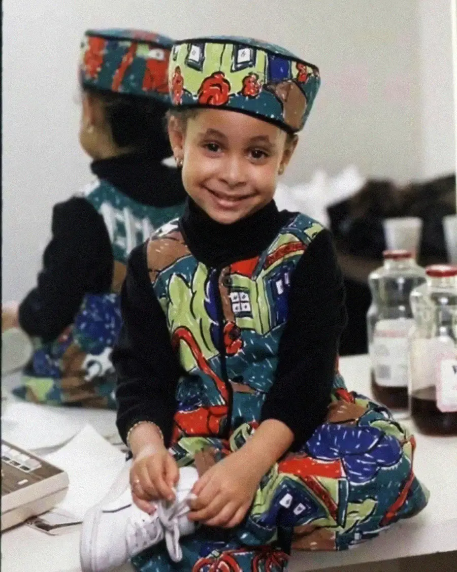 A young child sits on a counter, smiling at the camera. They wear a colorful, patterned outfit with a matching hat. In the background, their reflection is visible in a mirror, along with bottles and other objects.