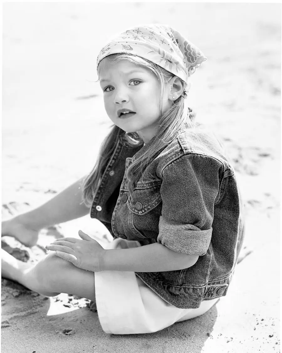 A young girl sits on the sand, wearing a denim jacket, a light-colored skirt, and a patterned bandana on her head, looking slightly to the side. The image is in black and white.