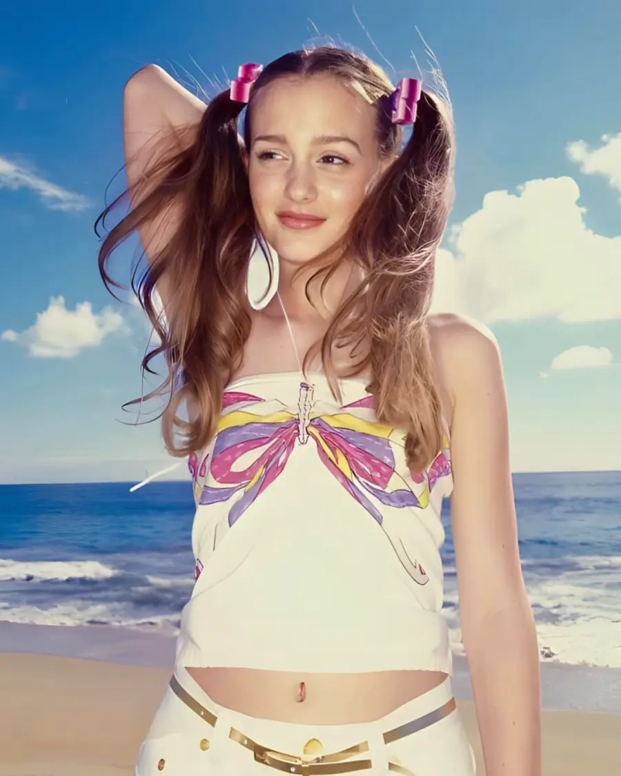 A young woman with long brown hair in pigtails, wearing pink hair clips, a butterfly-print tank top, and white pants, stands on a sunny beach with blue sky and clouds in the background.