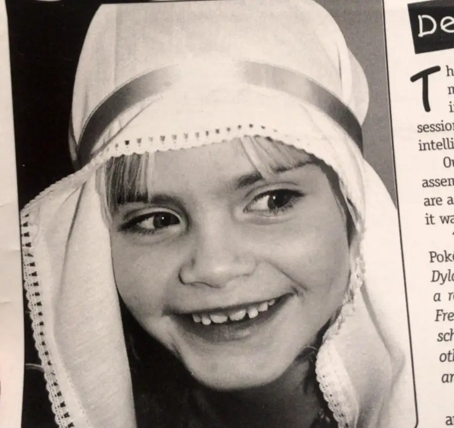 Black and white photo of a young child smiling, wearing a headpiece with a satin band and lace trim, possibly as part of a costume or play. The child has straight bangs and is looking slightly to the side.