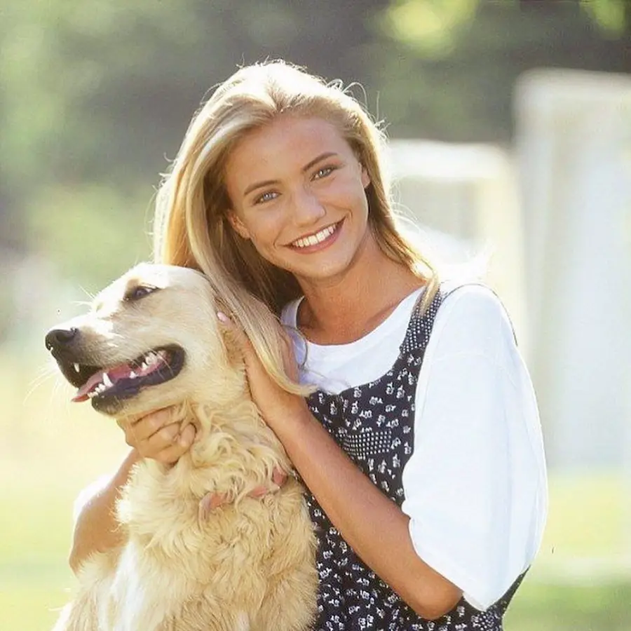 A young woman with long blonde hair smiles and hugs a golden retriever outdoors on a sunny day. She wears a white t-shirt under a patterned dress. The background is blurred greenery.