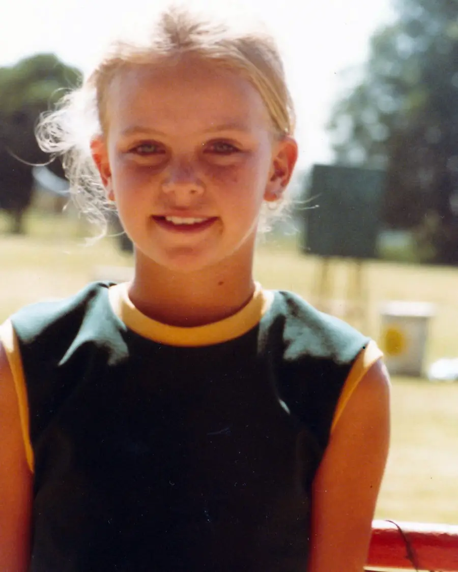 A young girl with blonde hair in a ponytail smiles at the camera. She is wearing a dark sleeveless shirt with yellow trim, standing outdoors on a sunny day with blurred trees and a field in the background.