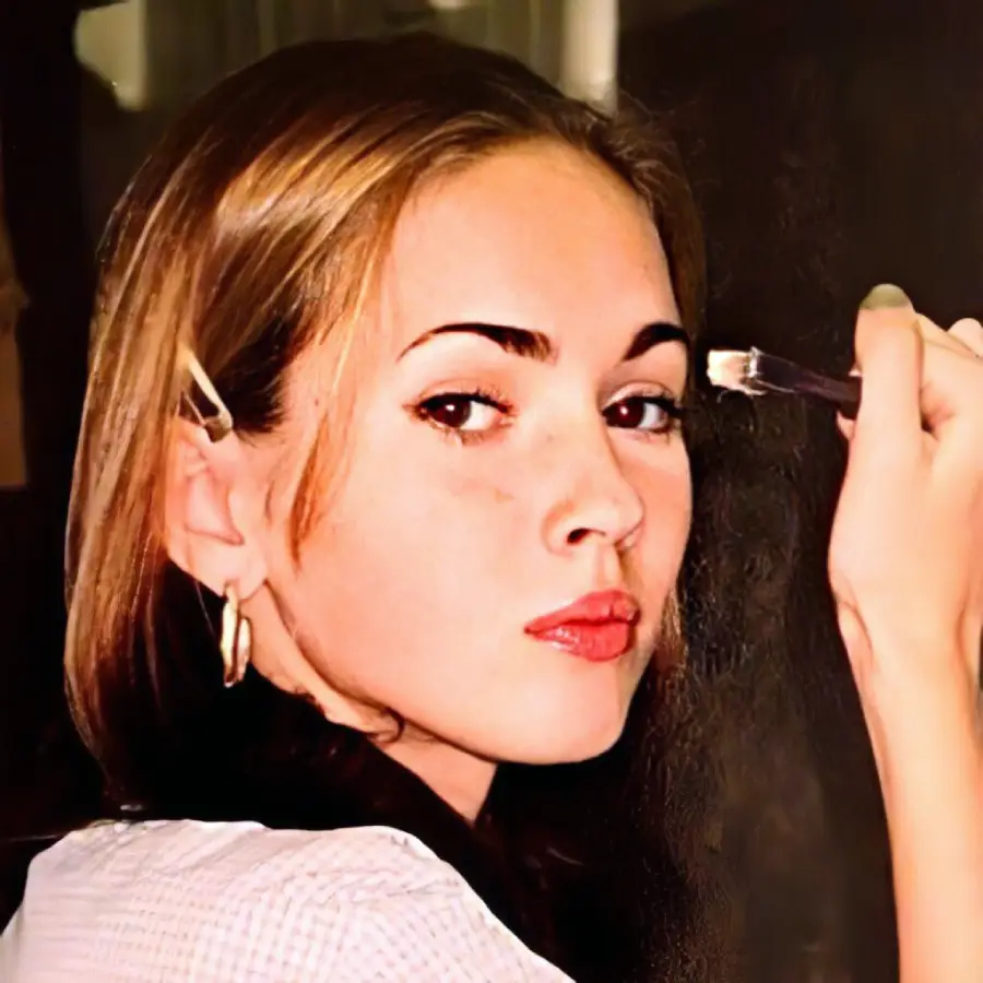 A young woman with light brown hair holds a makeup brush near her right eyebrow. She wears hoop earrings, a hair clip, and a checkered shirt, looking over her shoulder toward the camera with a neutral expression.