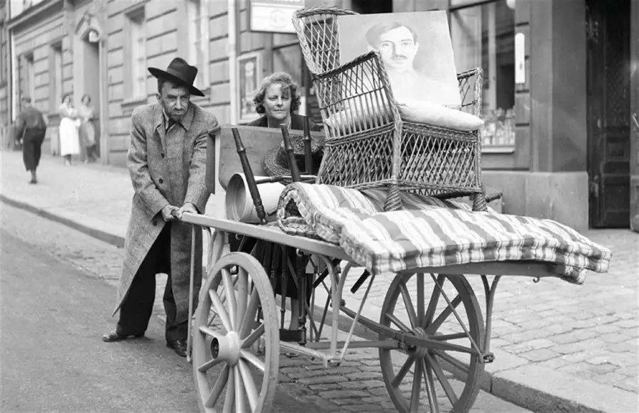 An elderly man pushes a woman in a cart with a large armchair and a portrait of a man on it, on a cobblestone street in an old city.