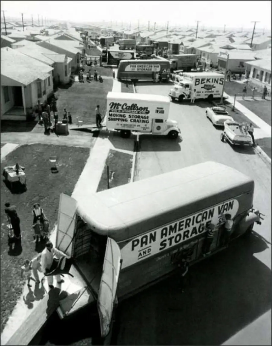 Black-and-white photo of a busy suburban street with multiple moving vans, people carrying boxes, and houses lining both sides; it appears many families are moving in at once.