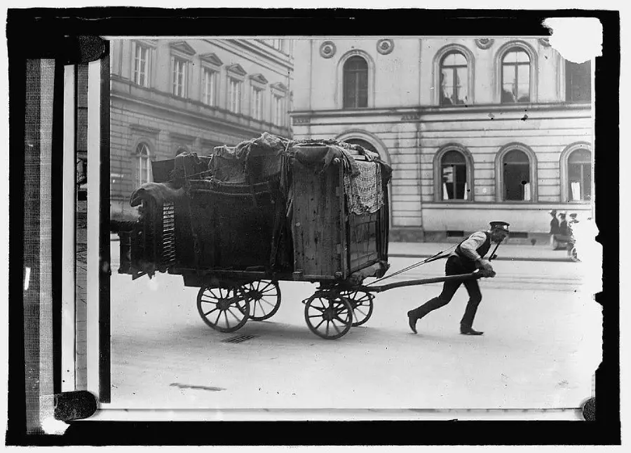 A man in uniform pulls a large wooden cart covered with fabric through a city street, with old buildings and a few pedestrians in the background.