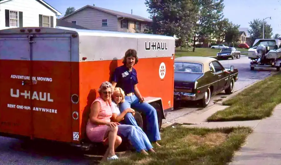 Three people sit on the back of a red and white U-Haul trailer parked on a suburban street, with houses, cars, and green lawns visible in the background.