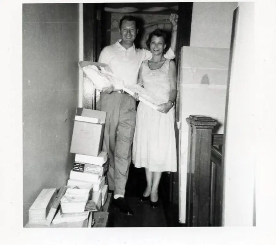 A smiling man and woman stand in a hallway holding a baby, surrounded by stacked boxes and a large wrapped item.