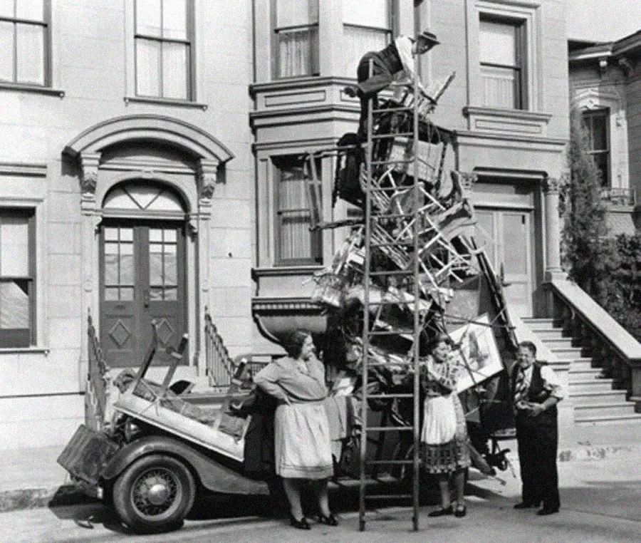Three people stand beside a car overloaded with household items stacked high above the roof. A tall, makeshift tower of furniture and belongings is precariously balanced, with a ladder leaning against it.