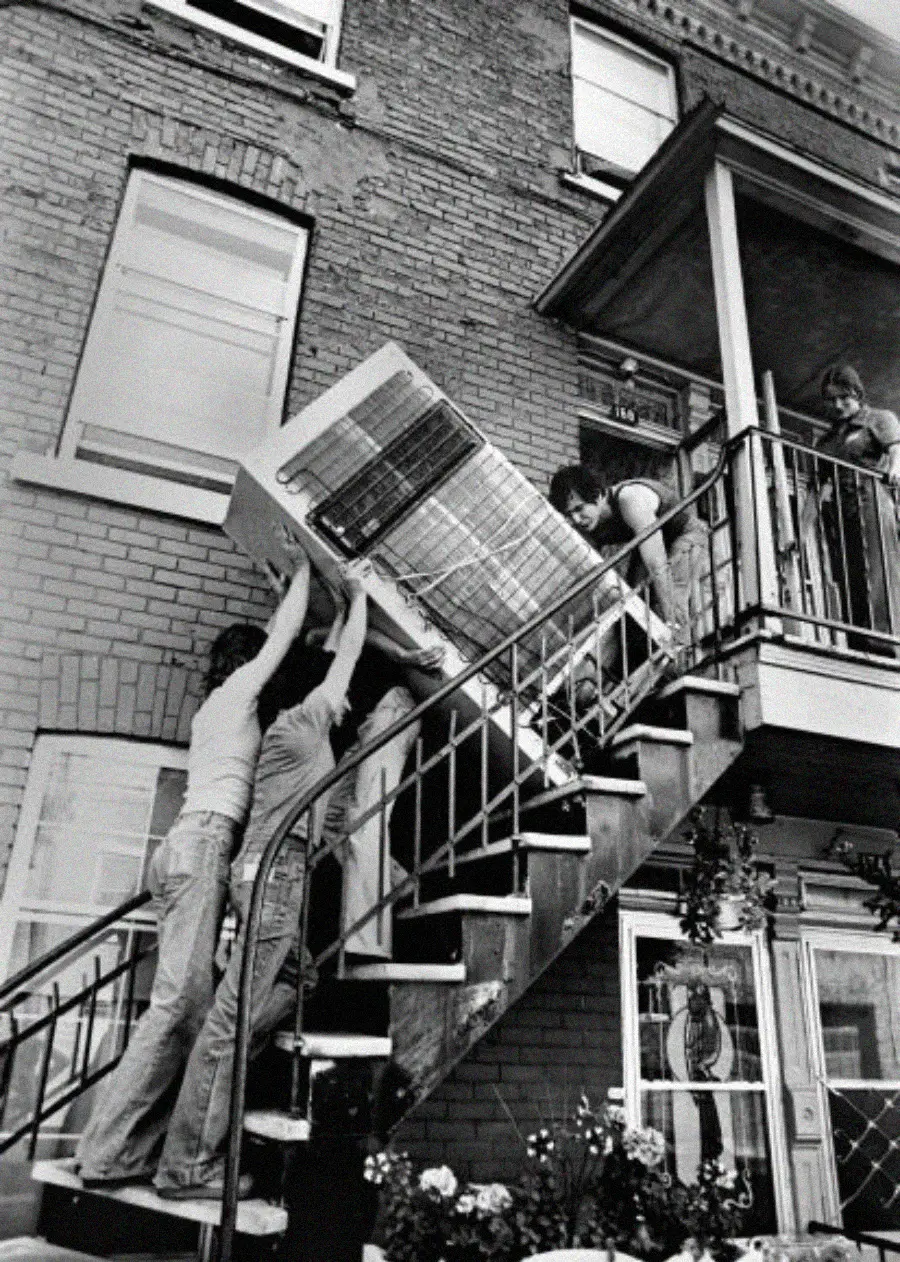 Four people struggle to lift a large couch up a narrow outdoor staircase toward a porch, while one person guides it from above. The scene takes place outside a brick building.