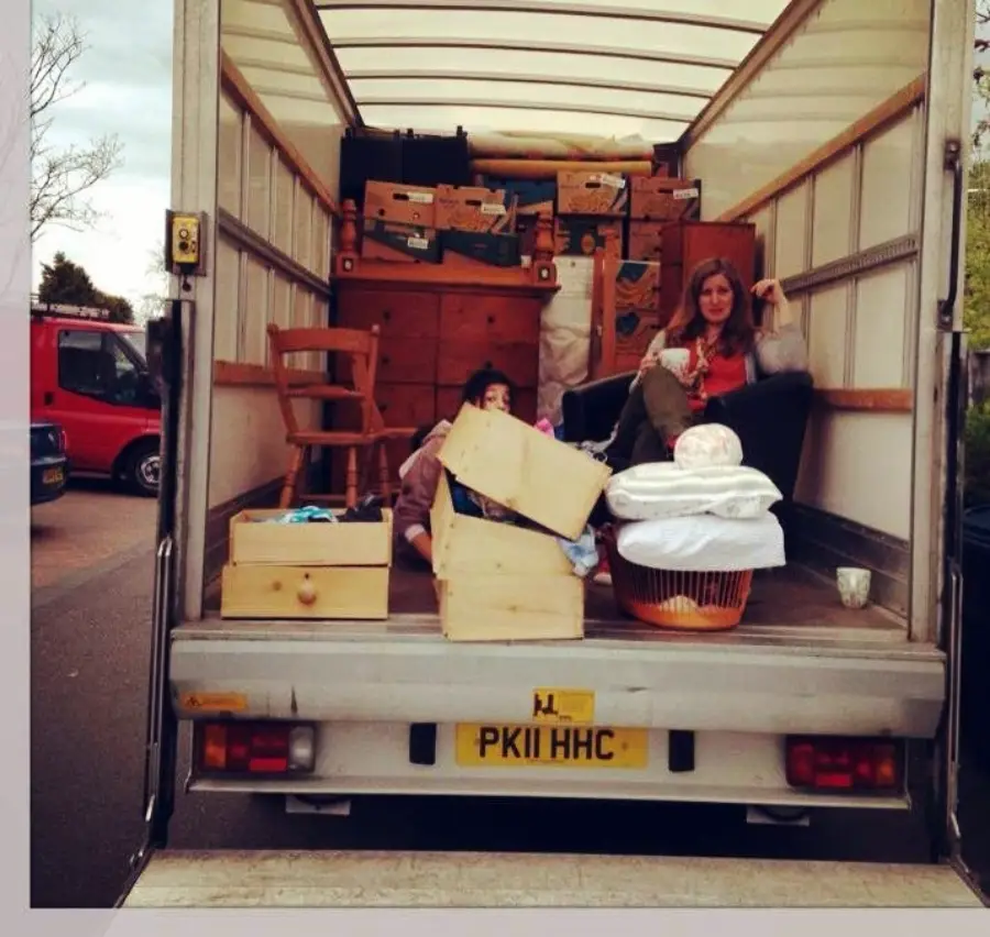 Two people sit among boxes and furniture inside the back of a moving truck. The truck is open, revealing packed items like chairs, baskets, and drawers. A red van is parked nearby on the left.