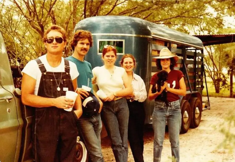 Five people stand beside a green truck and a trailer, outdoors on a sunny day. One person holds a drink, another holds a helmet, one has a puppy, and trees are in the background. All are smiling or relaxed.