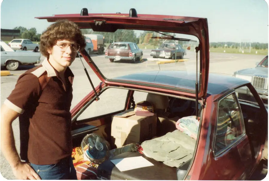 A young man with glasses stands by an open hatchback car trunk filled with boxes, clothes, and a fan in a sunny parking lot.