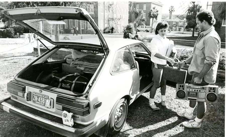 Two people stand by an open hatchback car, talking. The trunk is full of bags. One person holds a boombox. Trees and buildings are visible in the background. The scene appears to be from the 1980s.