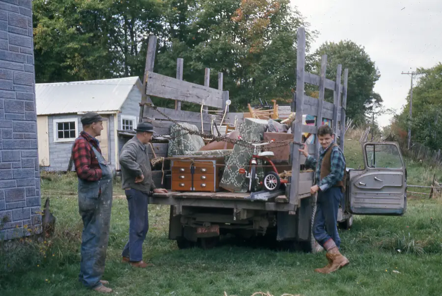 Three men unload furniture, chairs, and a tricycle from a flatbed truck beside a house on a grassy yard. Trees and another building are in the background.