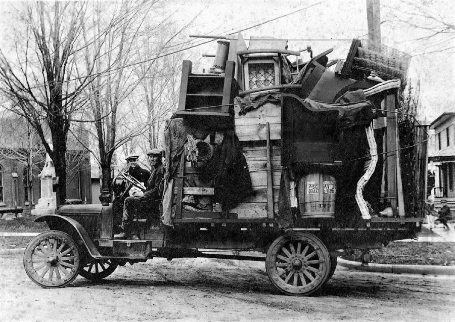 An old-fashioned truck overloaded with furniture, boxes, and household items is parked on a street. Two people sit in the cab. Leafless trees and houses appear in the background on a cloudy day.