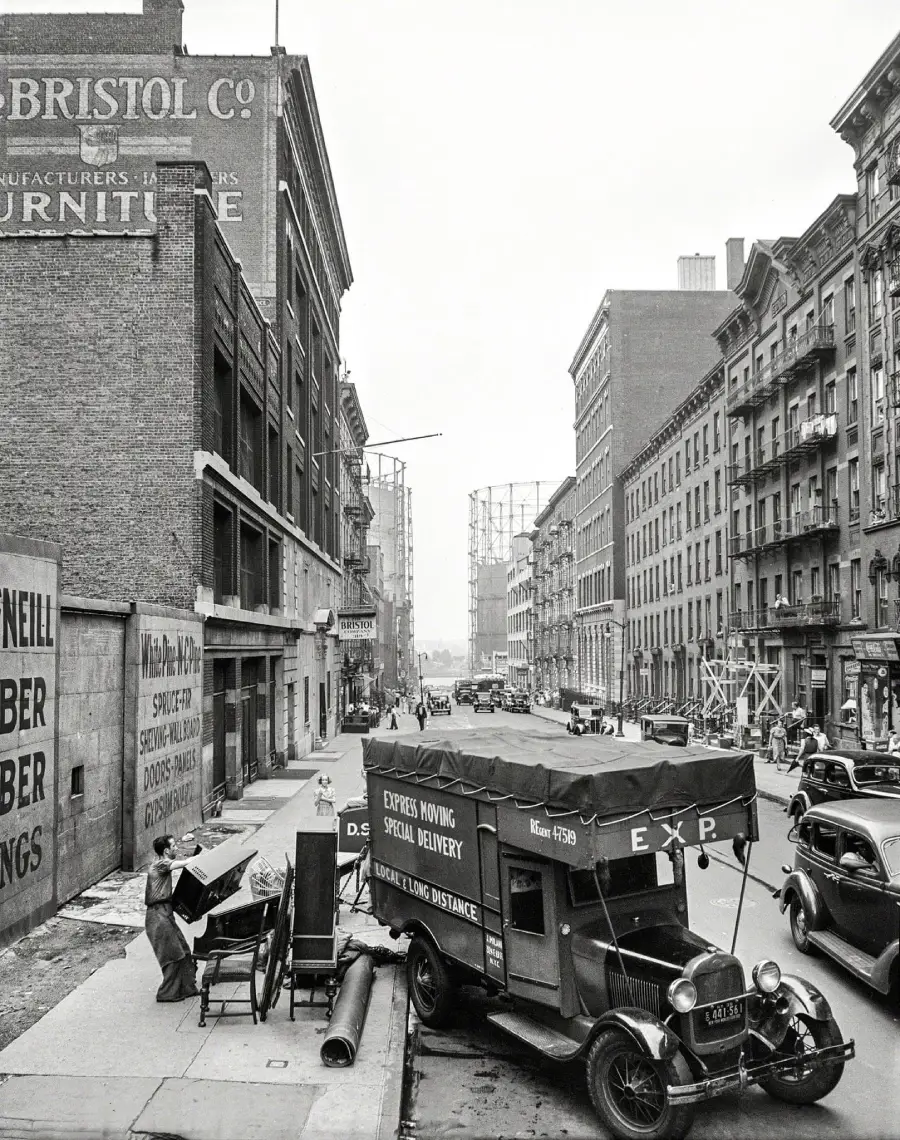 A black-and-white photo of a city street from the 1930s shows a moving truck parked curbside, with workers loading furniture. Old buildings and vintage cars line the street. Signs for furniture and moving services are visible.