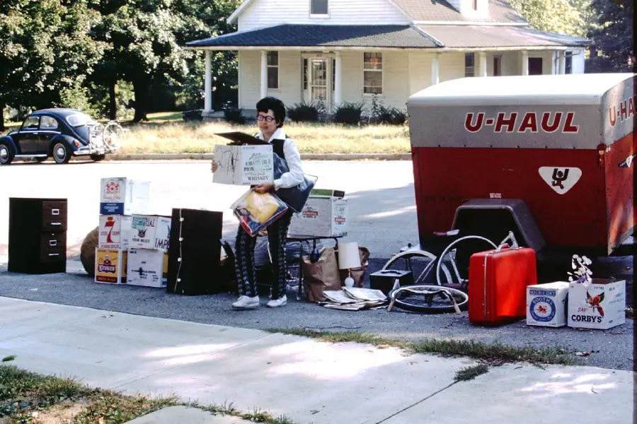 A person stands on a curb surrounded by boxes, suitcases, and furniture items next to a U-Haul trailer, preparing to move. A white house and a blue car are visible in the background.