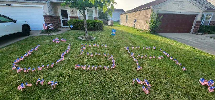 Dozens of small American flags arranged on a front lawn spell out "USA." There are more flags near the house, which has a tree and bushes and is next to a garage and driveway.