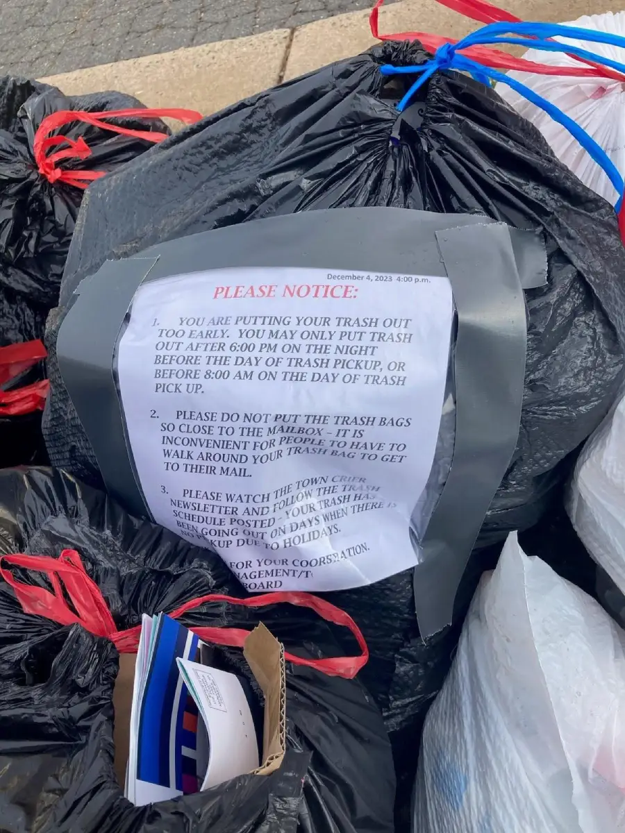 Close-up of a taped notice on a black garbage bag with red ties, surrounded by other trash bags and scattered mail. The notice warns about proper trash disposal times and requests not to block the mailbox.