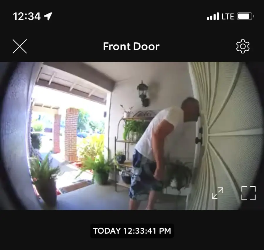 A man in a white shirt and shorts looks closely through a screen door, seen from a doorbell camera at a front porch with potted plants. Timestamp reads "TODAY 12:33:41 PM.