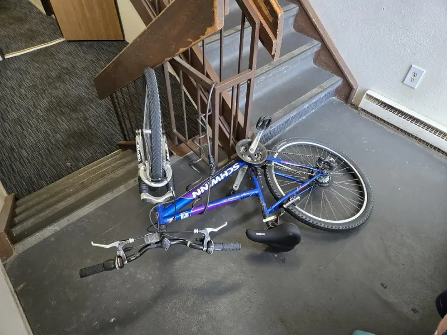 A blue Schwinn bicycle is lying on its side at the bottom of a stairwell, with the front wheel against the stairs and the seat detached on the floor nearby.