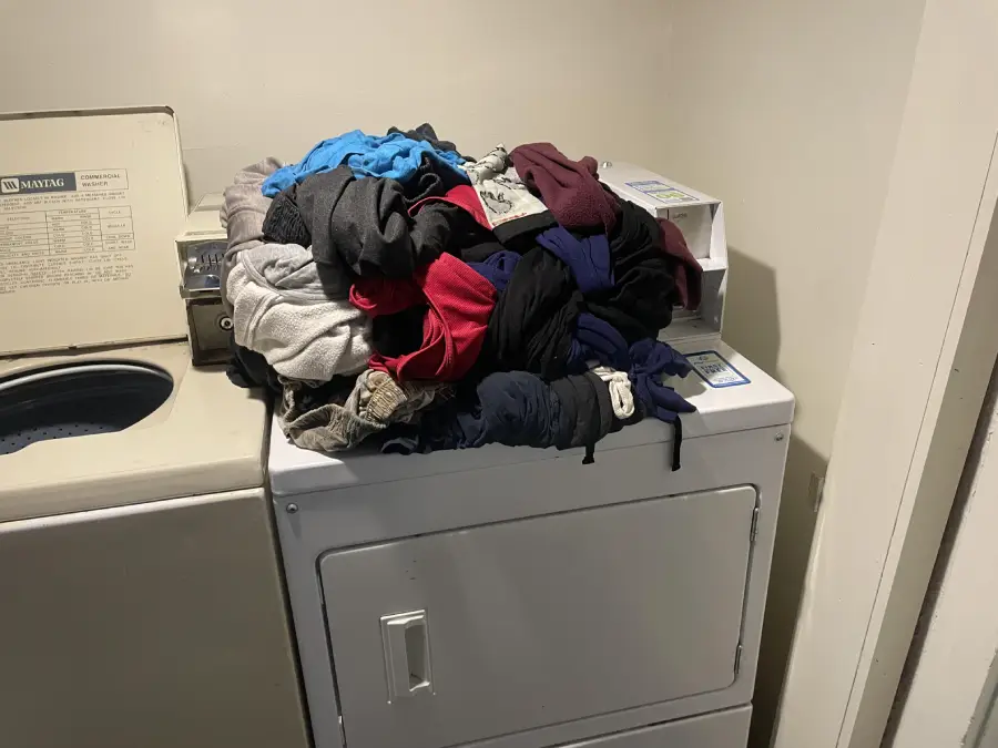 A large pile of assorted clothes sits on top of a white dryer next to an open washing machine in a laundry room with plain walls.