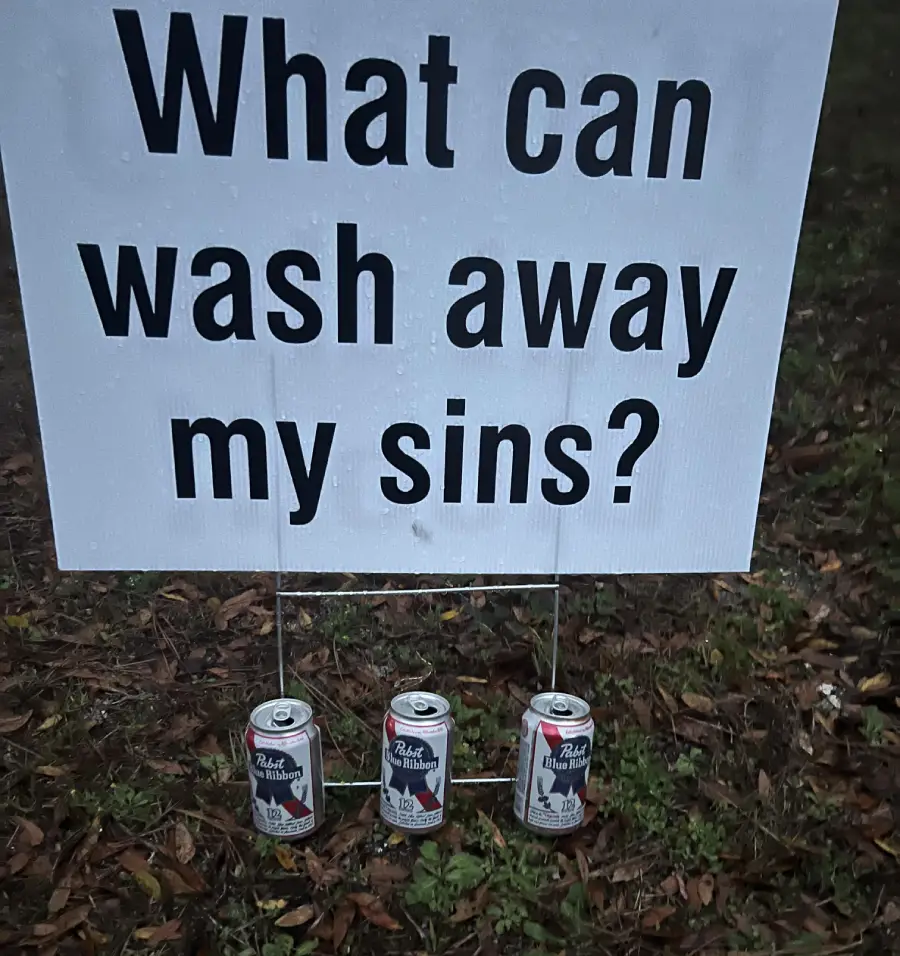 A sign reading "What can wash away my sins?" is displayed on grass, with three cans of Pabst Blue Ribbon beer placed underneath it.