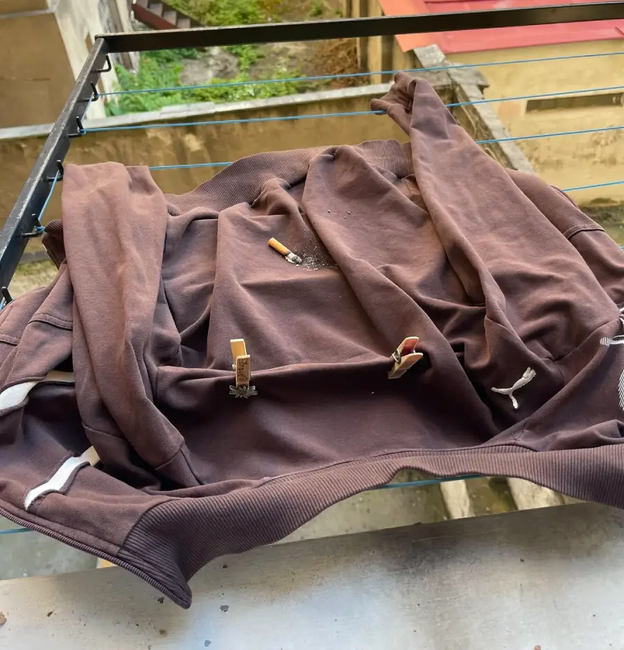 A brown sweatshirt is spread out and clipped to a metal drying rack with clothespins. The photo is taken from above, with buildings and greenery visible in the background.