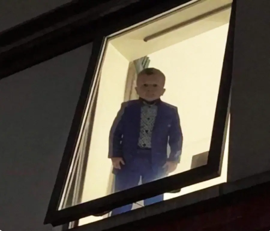 A young boy dressed in a blue suit and bow tie stands in a brightly lit room, looking out of an open, tilted window at night.