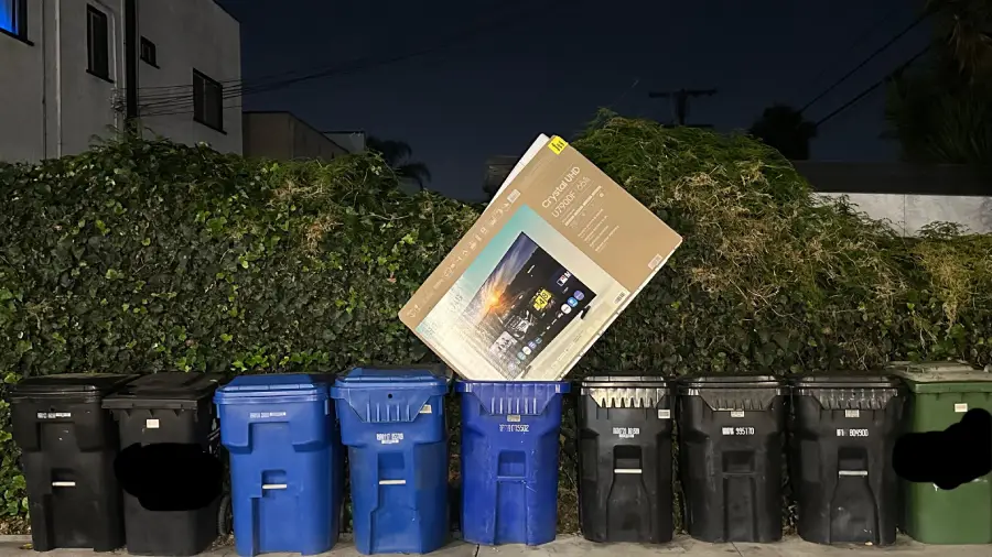 Eight garbage and recycling bins line a sidewalk at night, with a large, empty TV box leaning against the bushes behind them.