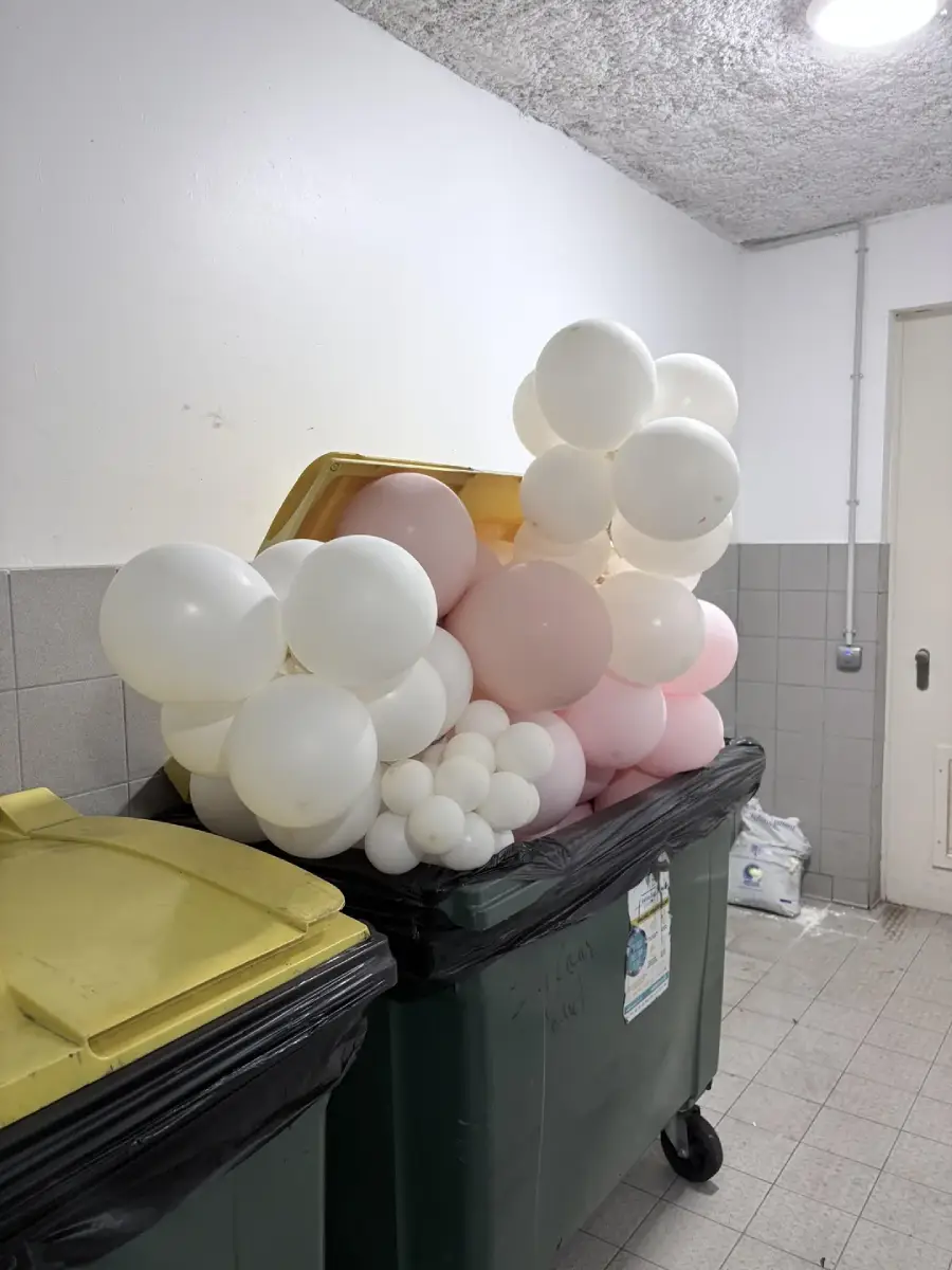 White and pink balloons overflowing from an open yellow-lidded dumpster in a tiled indoor area with another closed dumpster beside it.