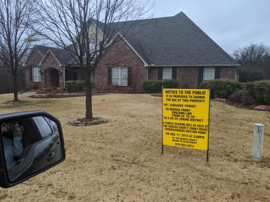 A brick house with a large yard has a yellow public notice sign about a proposed zoning change. A leafless tree stands nearby, and part of a car mirror is visible in the foreground.