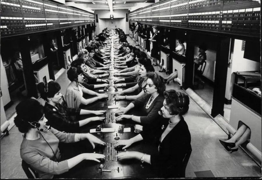 Black and white photo of rows of women working at a large telephone switchboard, wearing headsets and connecting calls, in a busy, vintage call center setting.