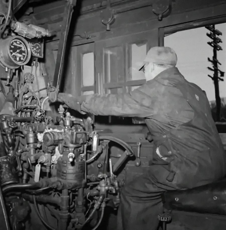 A man wearing work clothes operates controls inside a vintage train locomotive cab, surrounded by levers, gauges, and pipes, with a large window and telephone poles visible outside.