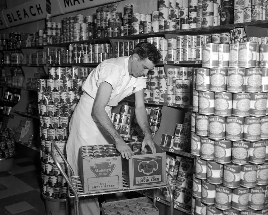 A grocery store worker in a white apron and shirt stocks shelves with canned goods, placing boxes of corn and green beans on a cart. Rows of canned food and cleaning products are visible on the shelves behind him.
