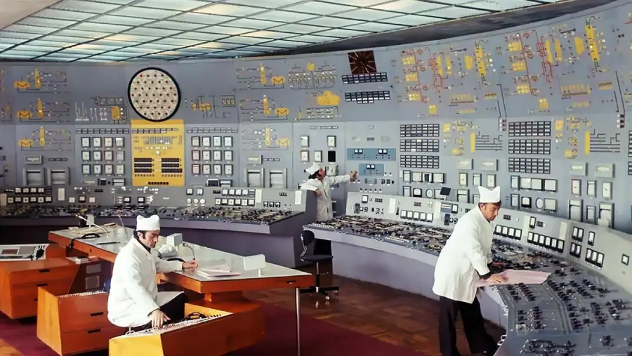 Three people in white uniforms work in a large, vintage control room with numerous buttons, dials, and displays on curved panels and walls. The ceiling has a grid pattern and the floor is wooden.