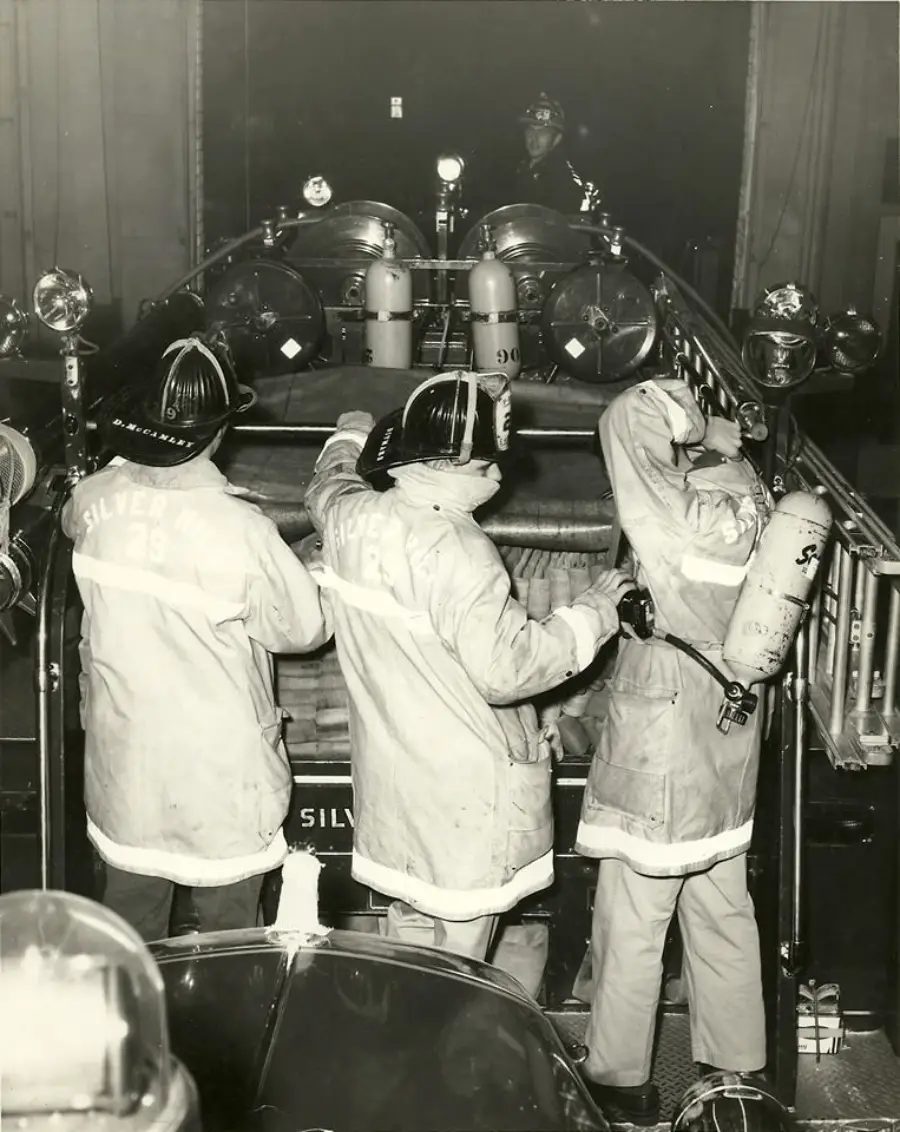 Three firefighters in gear stand at the back of a fire truck, preparing equipment. The truck has “Silverton” written on it and is equipped with hoses, lights, and tools.