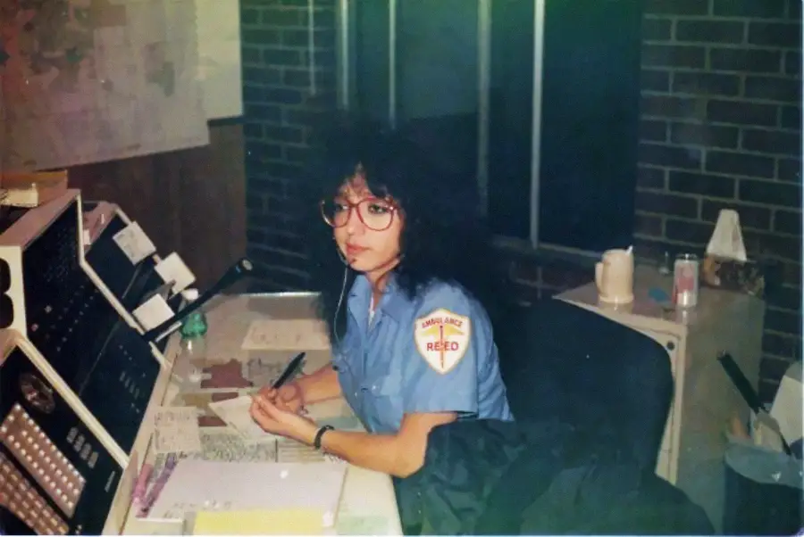 A woman with curly hair and glasses in a blue uniform sits at a desk with papers, a notepad, and communication equipment, inside an office with brick walls.