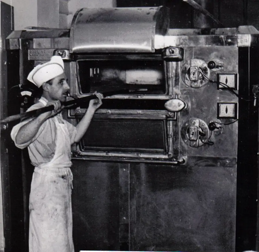 A baker in a white uniform and hat uses a wooden peel to place or remove bread from a large, industrial metal oven with dials and two doors. The scene appears to be from an old bakery.